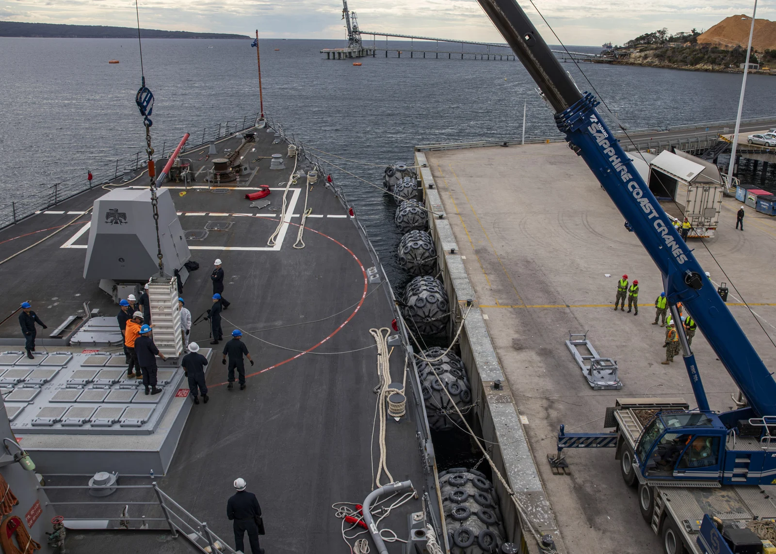 Une grue de quai descend un conteneur de missile sur le pont d'un navire de guerre gris amarré, entouré de marins casqués près des cellules de lancement