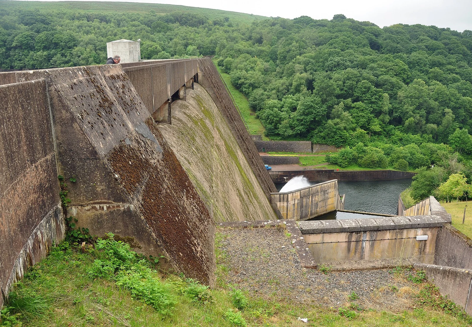 Le parement incliné en béton d'un barrage, avec un déversoir et un canal de restitution à son pied, parmi des collines boisées.