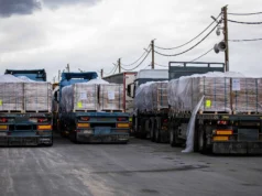 L’arithmétique d’une ration réduite A line of flatbed trucks loaded with aid pallets parked at a desert crossing point.