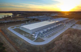 Who carries the debt behind the AI boom? Aerial view of a large, low grey data-centre building surrounded by rows of cooling equipment, with fields and a lake under a sunset sky.