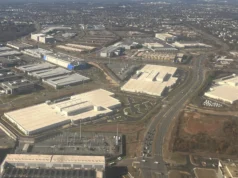 Qui paie le réseau dont les centres de données ont besoin ? Aerial view of large flat-roofed data centre buildings spread among roads and commercial sites under a partly cloudy sky.