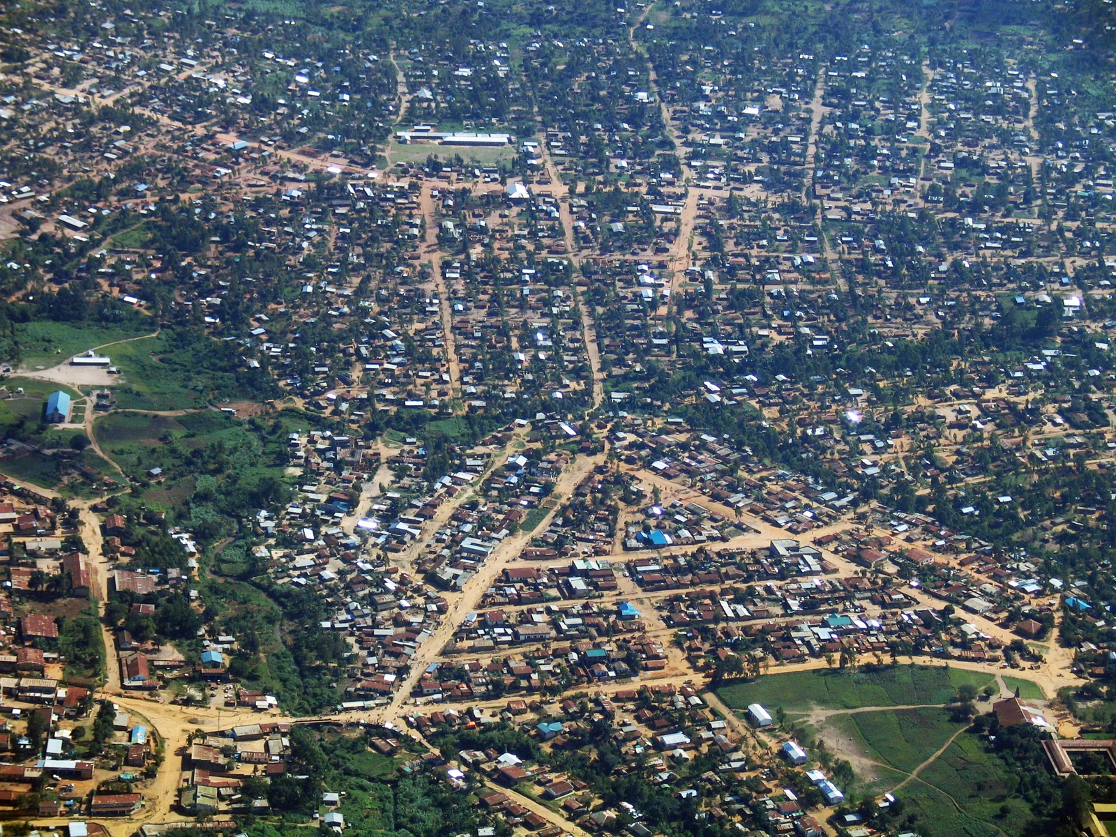 Vista aérea de una ciudad densa de casas pequeñas con tejados de chapa y caminos de tierra roja entre árboles verdes.