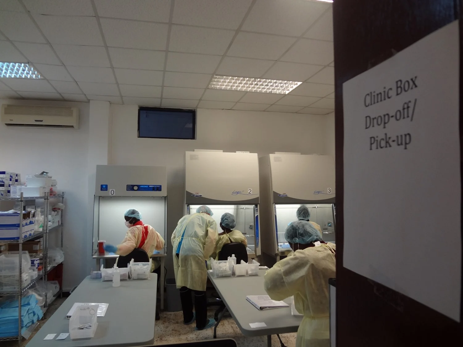 Farmacéuticos con batas y gorros de protección trabajan en mesas de laboratorio y cabinas de seguridad en una sala de una clínica.