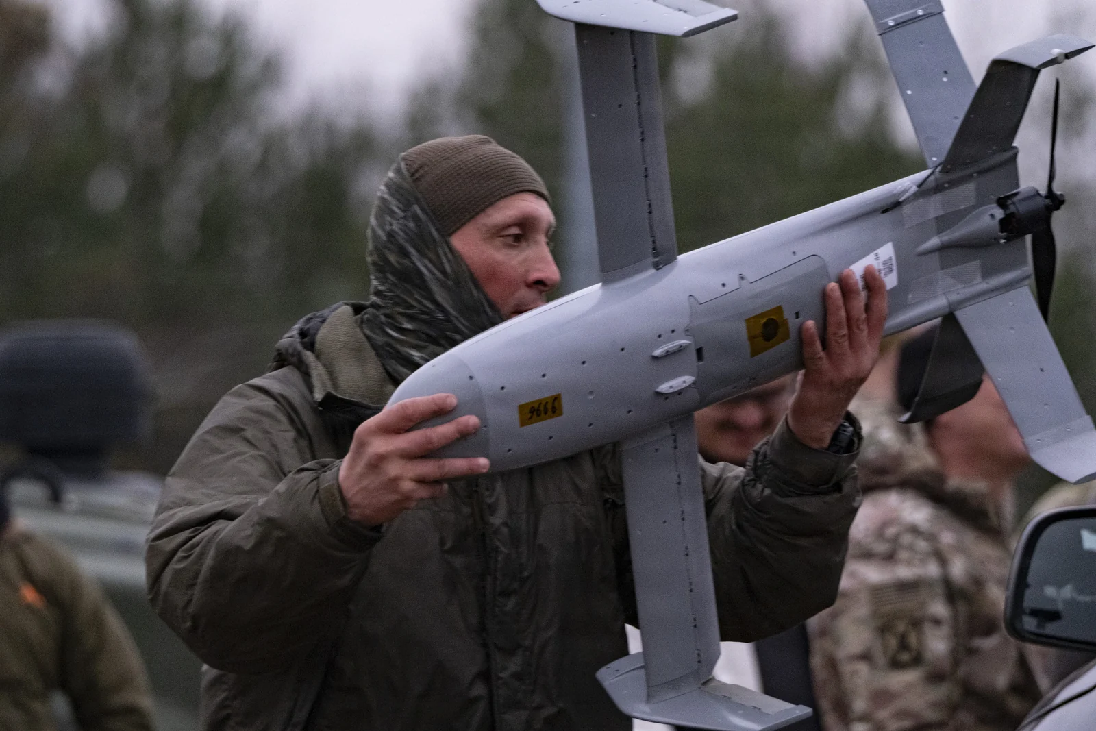 Un soldado con gorro de punto y bufanda levanta un pequeño dron gris de ala fija con hélice trasera