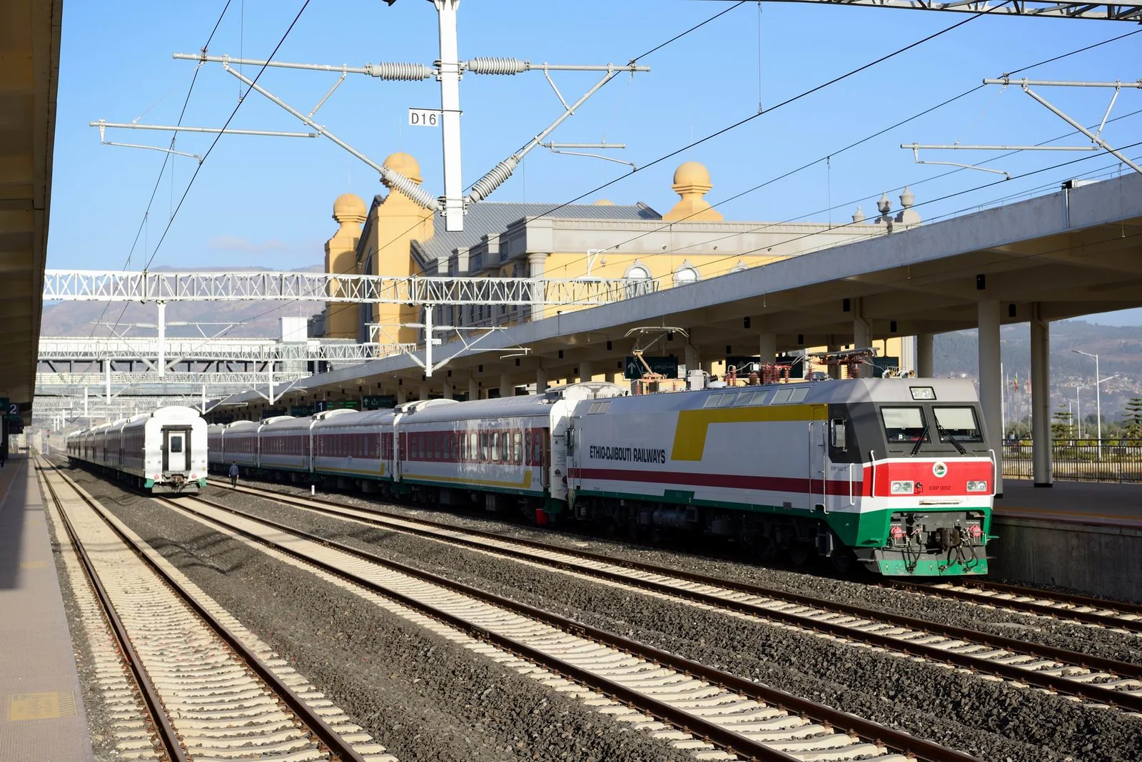 A green, white and red electric locomotive hauling white passenger coaches under overhead wires at a modern railway station