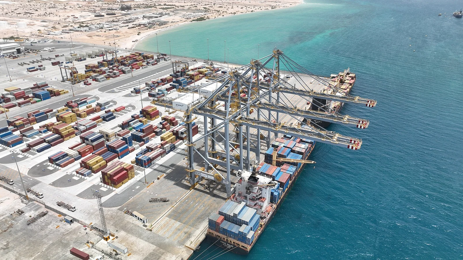 Aerial view of a container terminal with ship-to-shore cranes, stacked containers and a moored container ship beside turquoise water