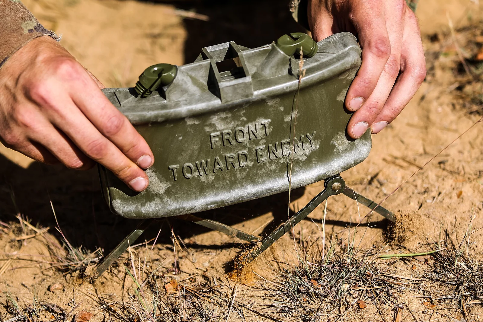 Dos manos colocan una mina Claymore verde y curvada, con la inscripción «Front toward enemy», sobre sus patas plegables en un suelo arenoso
