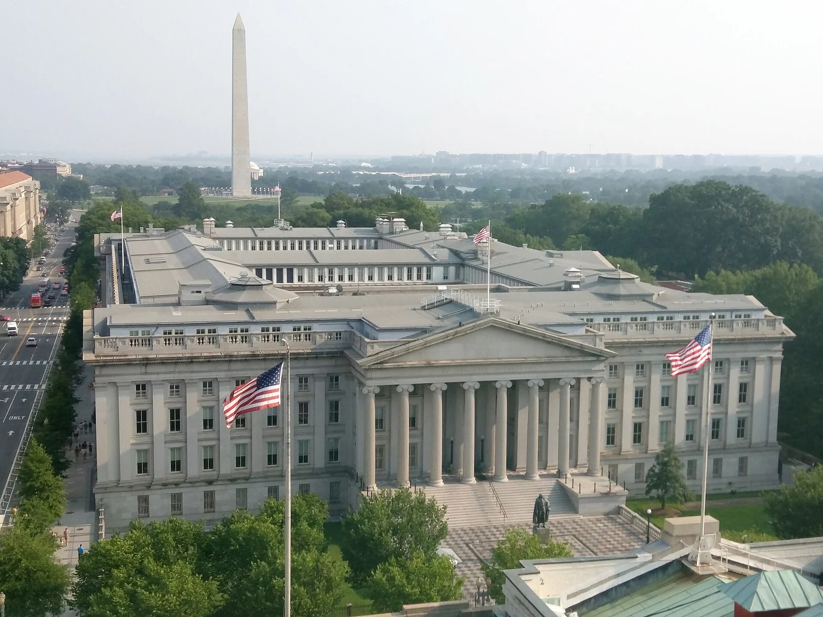 Luftaufnahme des klassizistischen Gebäudes des US-Finanzministeriums mit amerikanischen Flaggen davor und dem in der Ferne aufragenden Washington Monument.