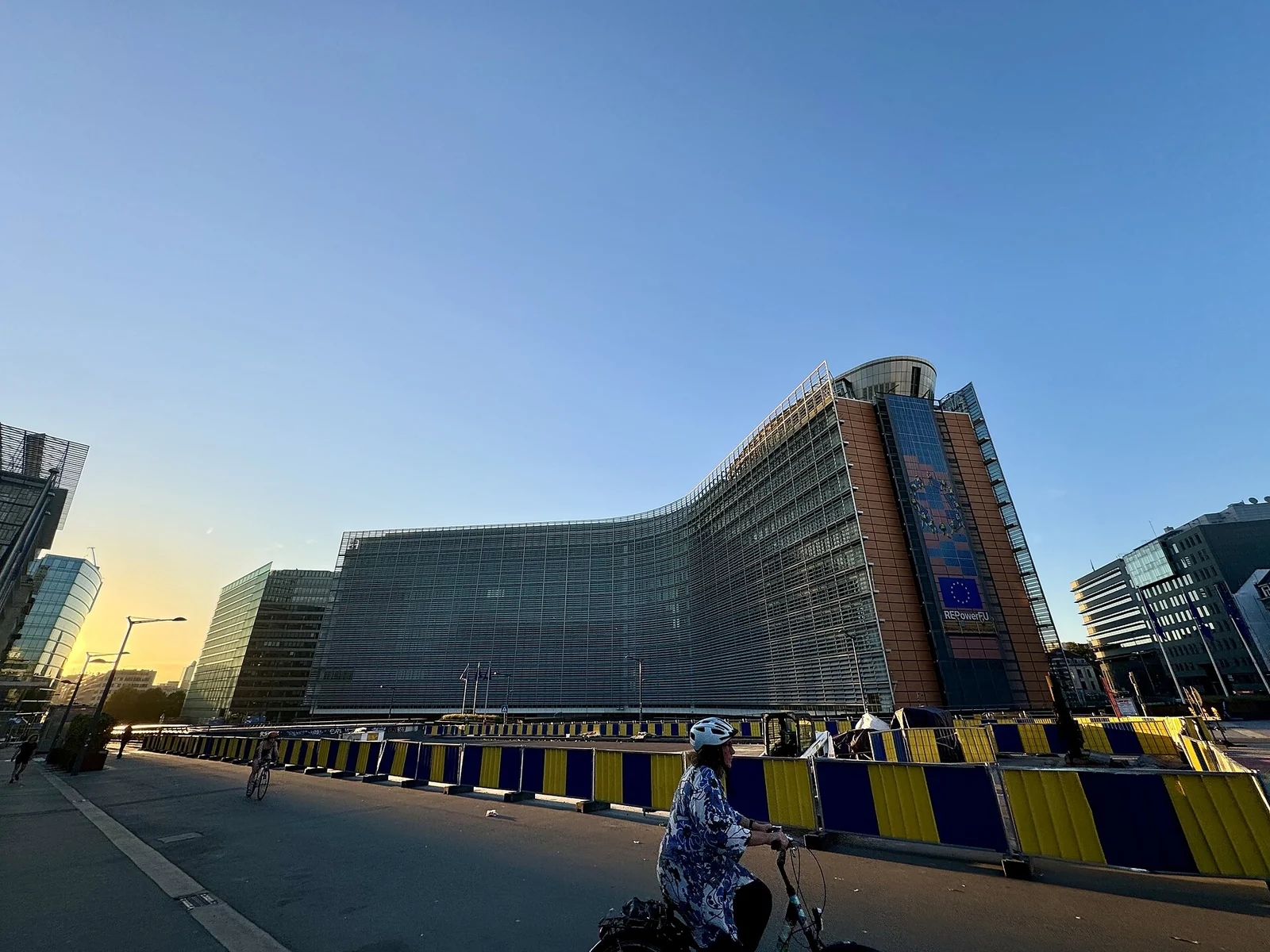 Die geschwungene Glasfassade des Berlaymont-Gebäudes in Brüssel in der Abenddämmerung, mit EU-Flaggen am Gebäude und blau-gelben Absperrgittern entlang der Straße.