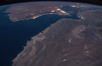 How the world absorbed the Hormuz oil shock Night view from orbit of the Gulf of Oman and the Strait of Hormuz, with city lights along the coasts and the curve of the Earth on the horizon
