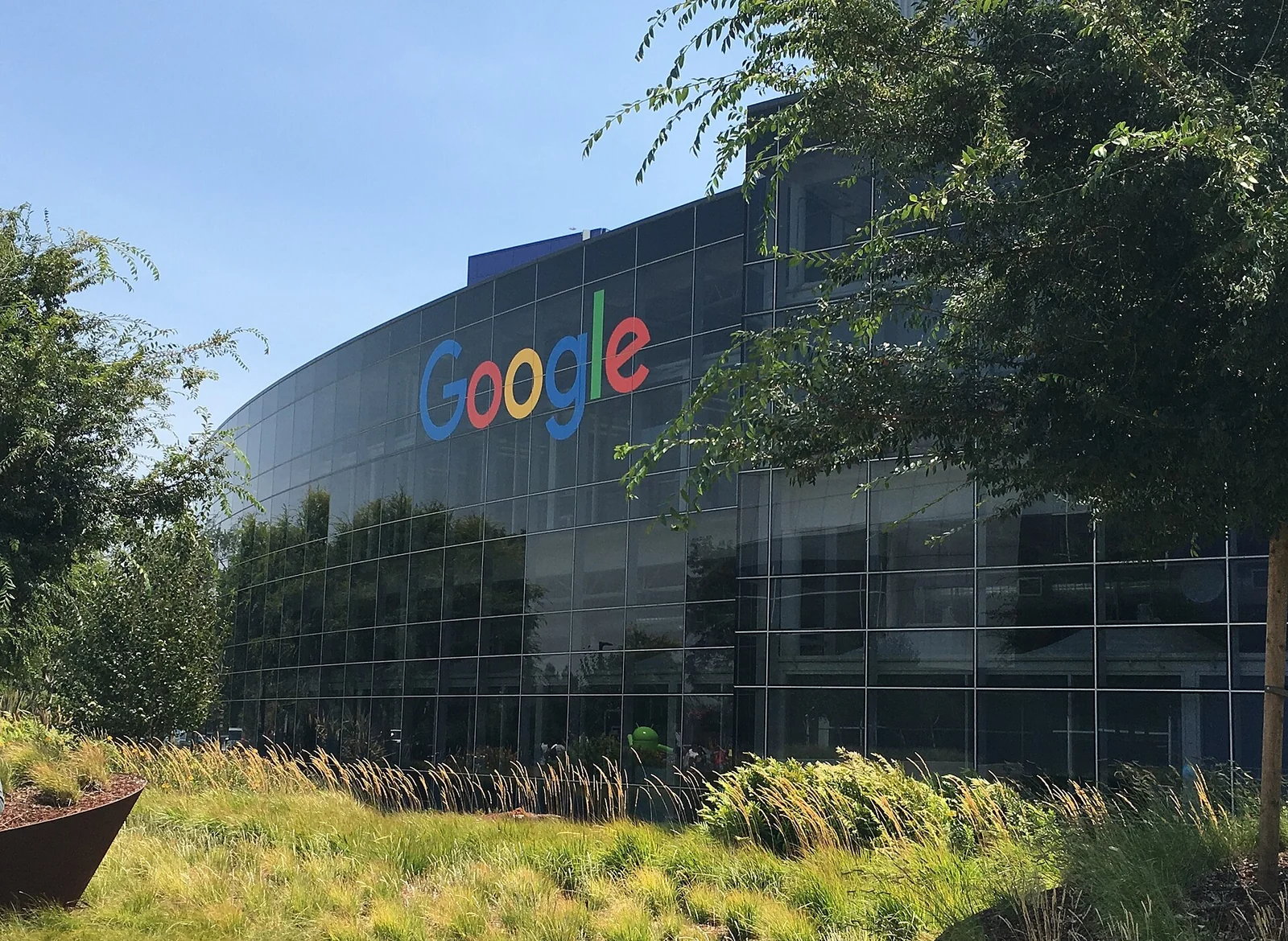 Un edificio de oficinas curvo de cristal con el logotipo multicolor de Google en la fachada, detrás de árboles y césped.