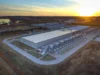 AIブームを支える債務は誰が背負うのか Aerial view of a large, low grey data-centre building surrounded by rows of cooling equipment, with fields and a lake under a sunset sky.