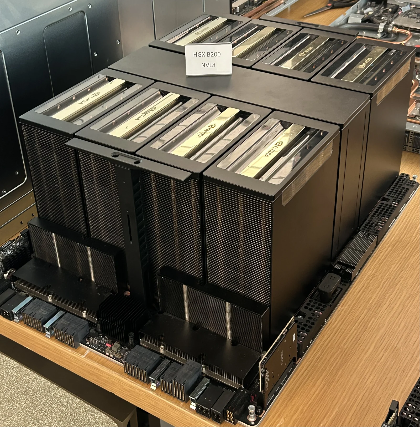 Una placa de servidor con ocho grandes módulos de GPU negros cubiertos por disipadores, expuesta sobre una mesa de madera.