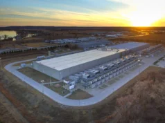 ¿Quién carga con la deuda detrás del auge de la IA? Aerial view of a large, low grey data-centre building surrounded by rows of cooling equipment, with fields and a lake under a sunset sky.