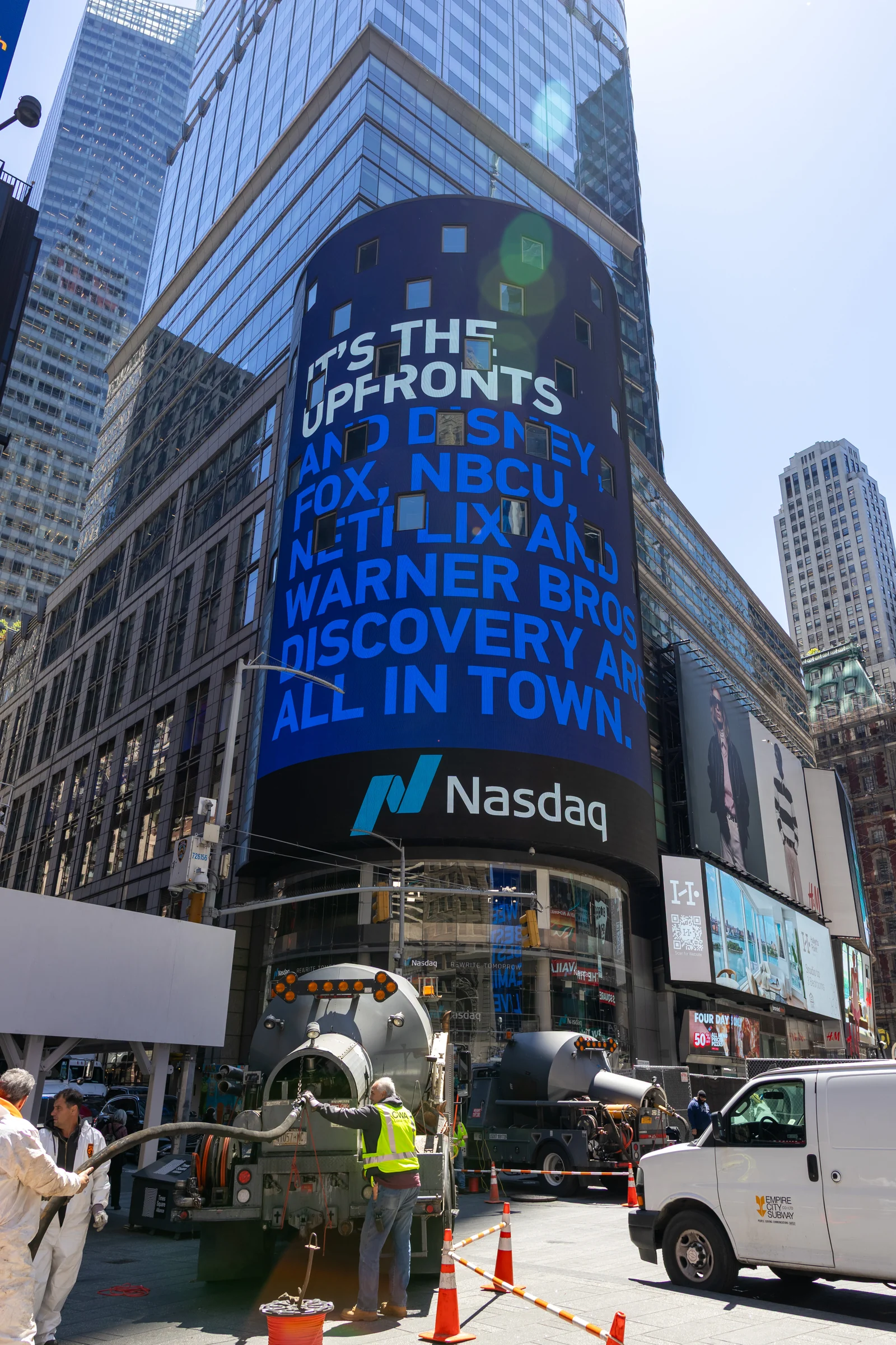 La pantalla cilíndrica de vídeo del Nasdaq en un rascacielos de Times Square, con el logotipo del Nasdaq, sobre una calle con obreros de la construcción.