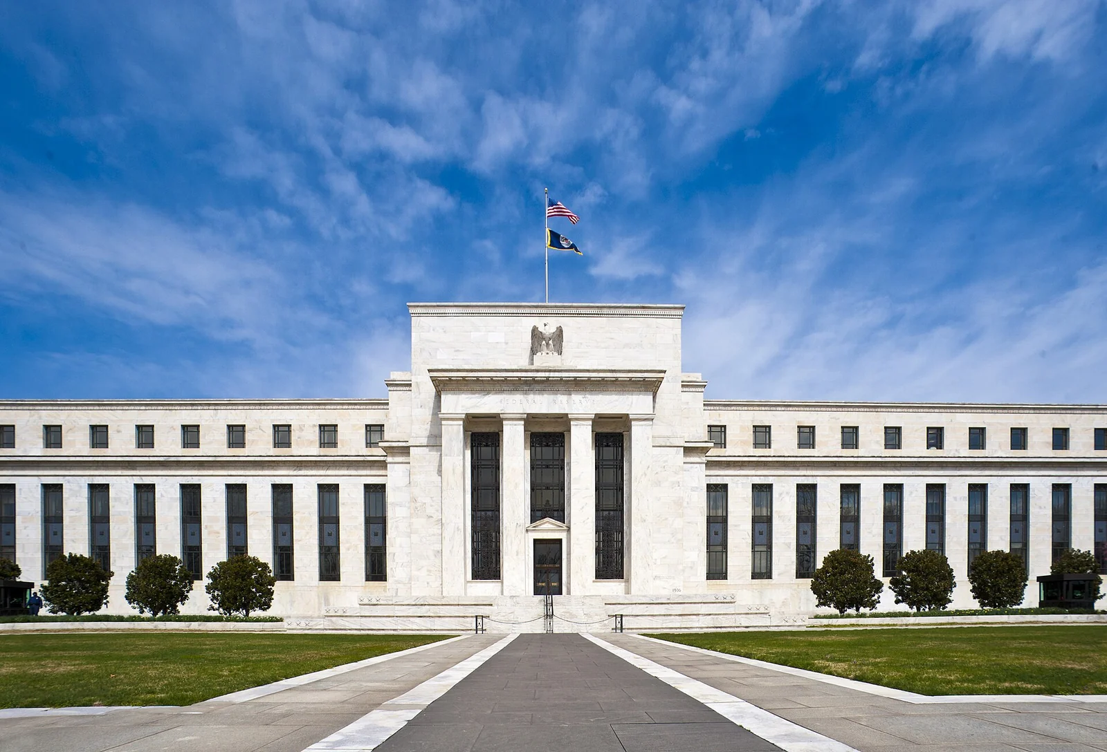 Symmetrical white marble facade of the Federal Reserve Eccles Building behind a lawn and a central walkway, under a blue sky with clouds